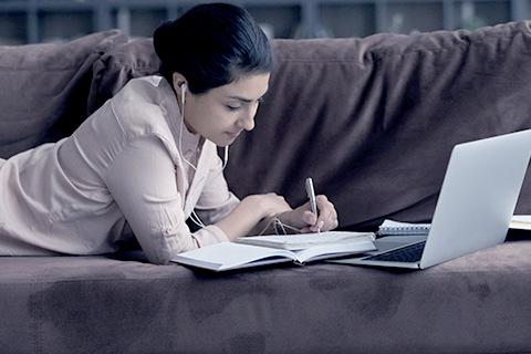 Girl lay on couch, writing in notebook with laptop in front of her